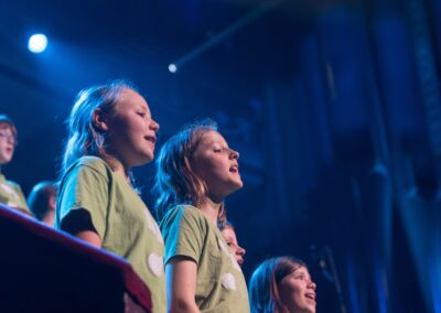Several girls in green LYC T shirts, singing, with more singers visible behind them, against a background lit in dark blue.