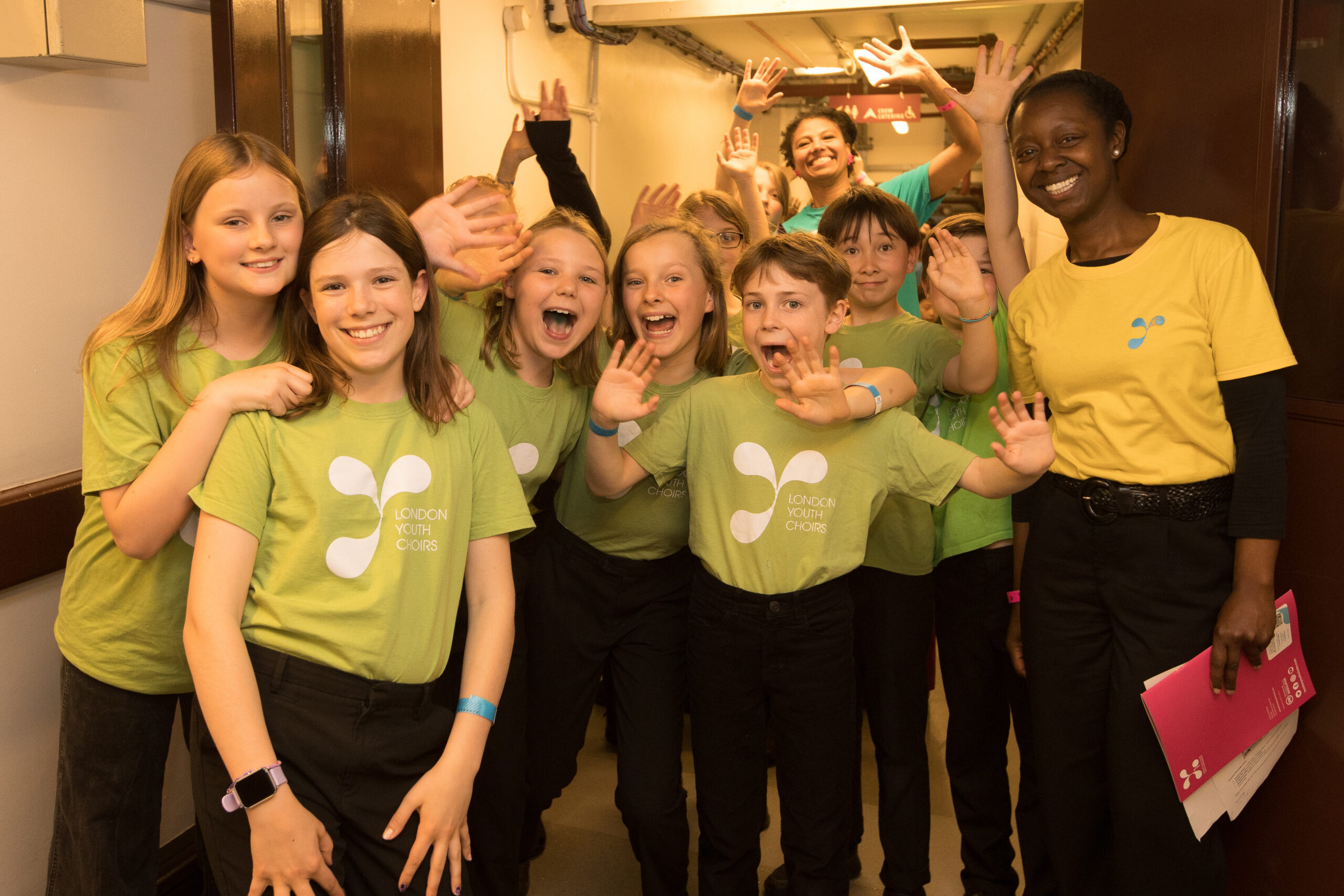 A group of LYC South East members age 7-11 wearing green LYC T shirts and black trousers, with a staff member in a yellow T shirt and another staff member in the background, smiling and making silly faces at the camera backstage at the royal albert hall