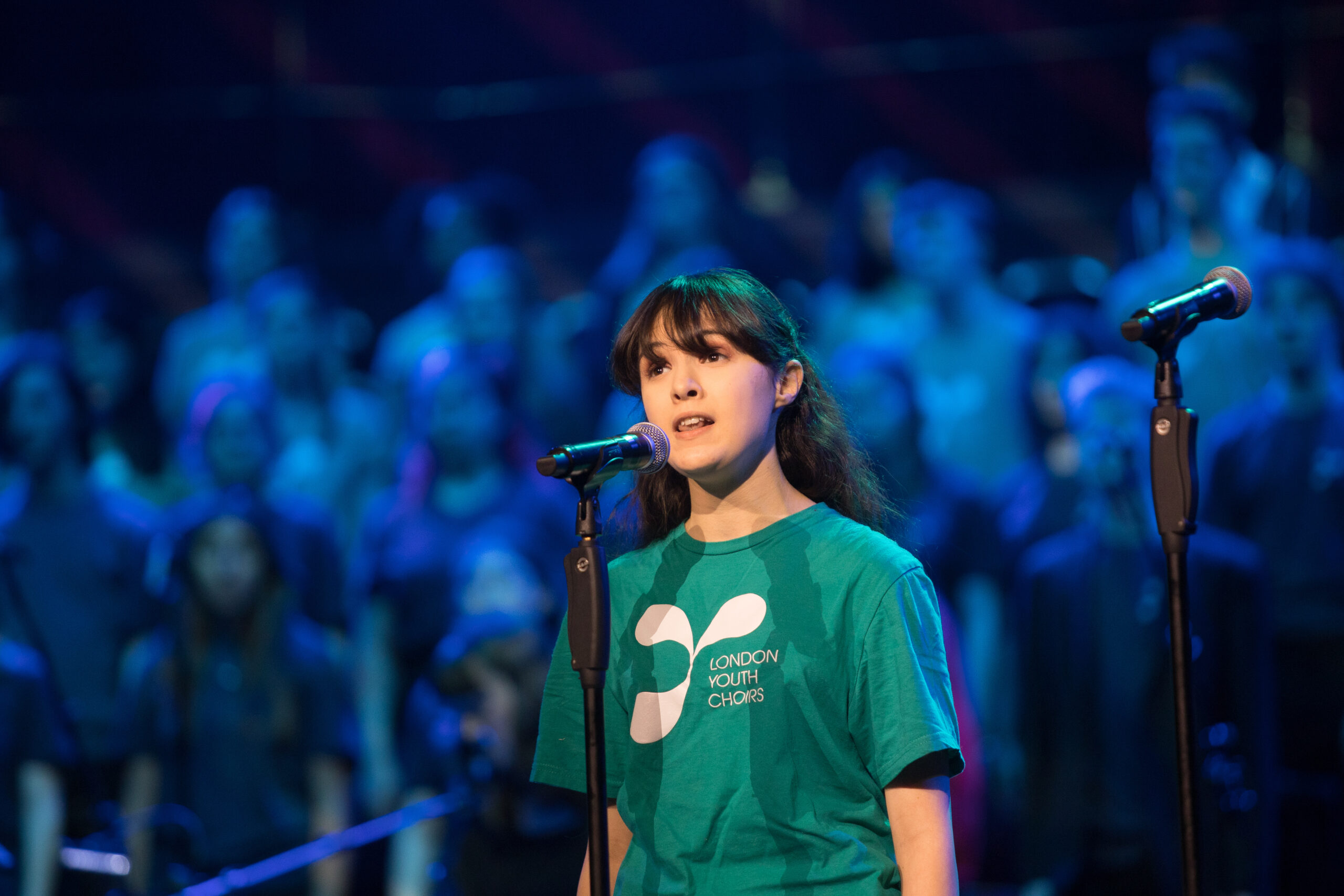 LYC member Grace wearing a teal T shirt with white LYC logo, singing into a microphone, with a choir behind under blue lighting