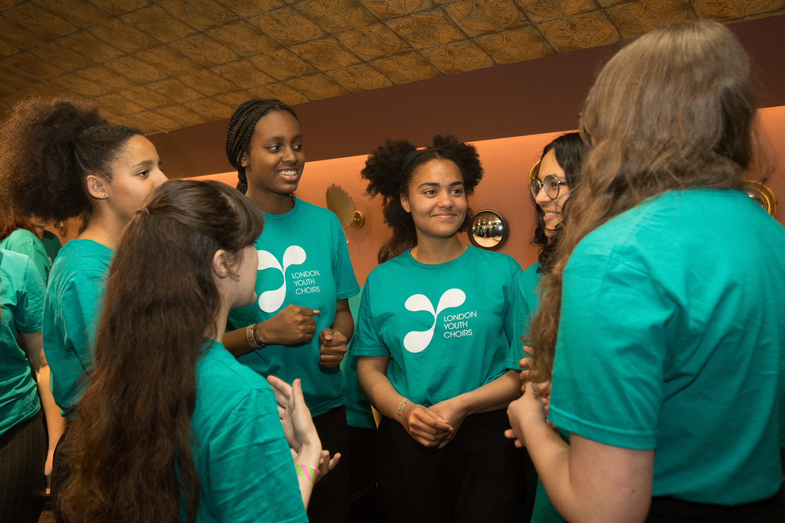 A group of 6 LYC members, age 16-23 and female, stood in a circle chatting, wearing teal LYC T shirts