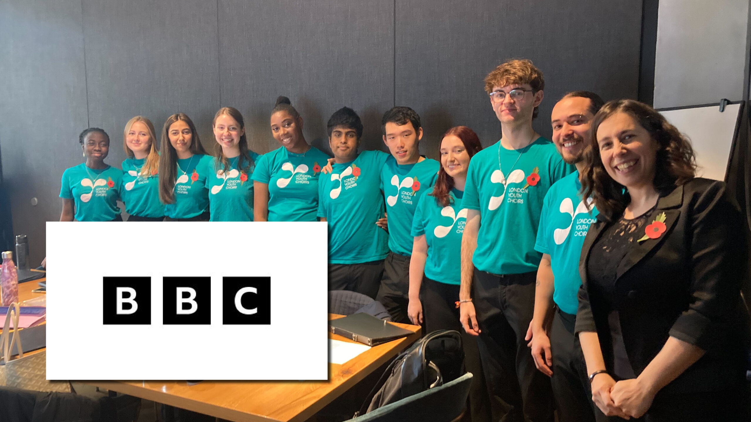 10 Chamber Choir members in Teal T shirts, wearing poppies, with Rachel Staunton on the right dressed in black with a poppy. A black and white BBC logo is in the bottom left of the image.