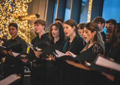 5 members of LYC Chamber choir in a row, wearing smart black concert attire, holding black folders and singing. At the back of the singers on the left of the image is a lit up Christmas tree. Behind are floor to ceiling windows with a dark sky visible.