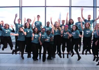 LYC Chamber Choir wearing teal LYC t shirts and black trousers, jumping up with their arms in the air and smiling at the camera. The back drop is floor to ceiling windows.