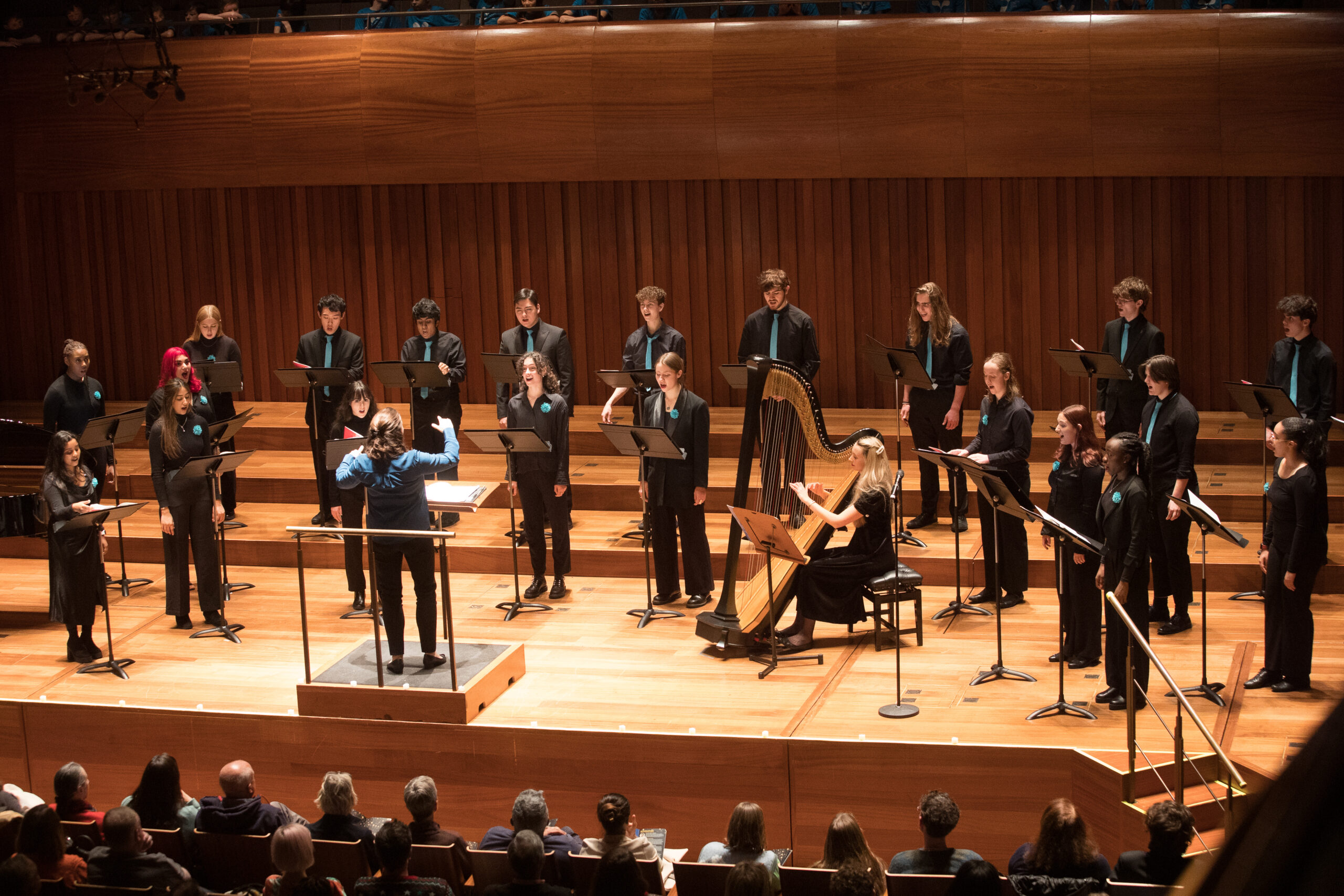 LYC Chamber Choir standing in two rows on a concert hall stage, each with a music stand. They are wearing black concert dress with teal accessories. The conductor is in front of them wearing a blue blouse and a harpist is sitting to the right of the conductor.