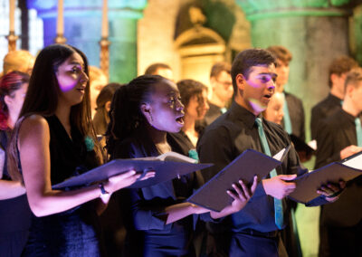4 chamber choir members wearing concert black with a teal brooch or tie, holding black folders and singing, with another row of singers visible behind. the singers are in a church which is lit in blue, green and yellow behind them.