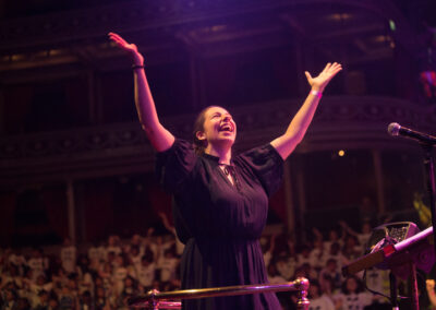 A woman with hair tied back, wearing a black dress, with her arms up in the air and with a joyful expression. In the background, rows of children in white T shirts are visible