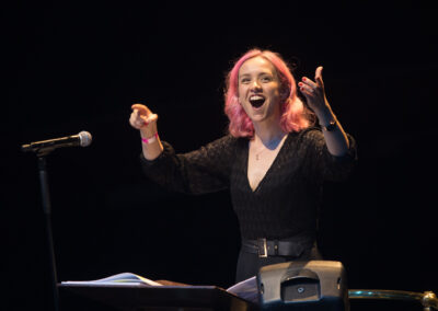 A woman with pink hair in a black jumpsuit, conducting against a black background