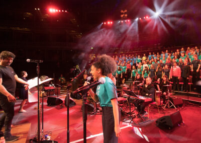 Red lighting, and a girl stands in the centre of the picture, singing into a microphone. To the left is the conductor, and to the right behind the soloist is a band and about 6 rows of singers in LYC T shirts.