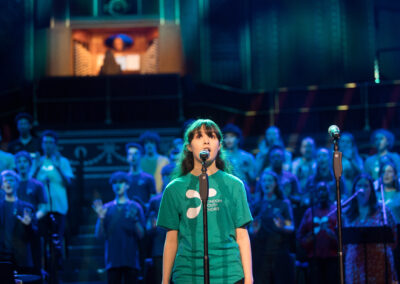 A girl standing at a microphone and wearing a T shirt with an LYC logo. behind her, the rest of the choir is visible in blue lighting, with the organ seen above.