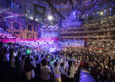 The Royal Albert Hall, with the stage on the left and audience on the right. Spotlights light up the stage in blue, and young people in LYC T shirts are on the stage, in the choir stalls, and in the right and left most audience seats. The audience is shining their phone torches, and the performers are holding finger torches.