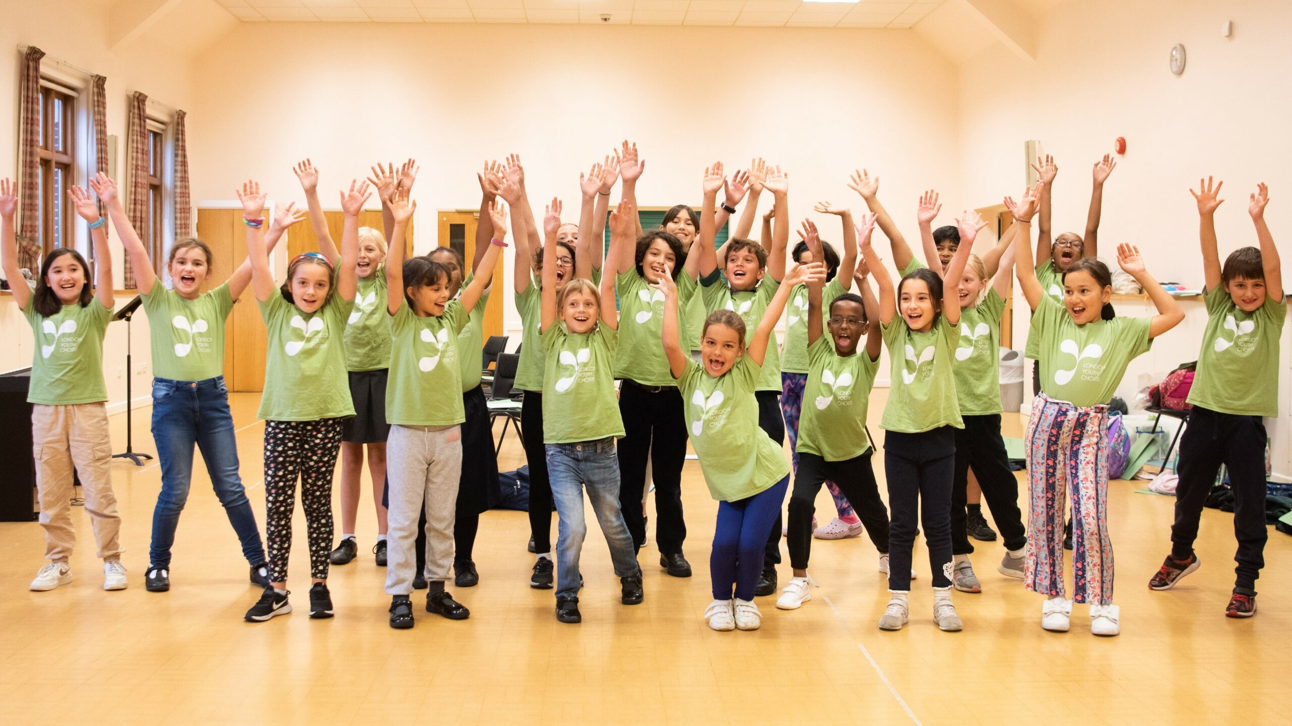 About 20 children wearing green LYC T shirts, with their arms in the air, in a beige coloured hall.