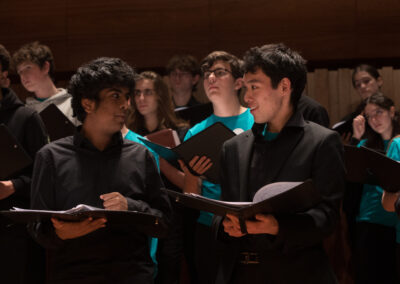 Two young men dressed in black, holding choir folders and talking to each other. Behind, more singers are visible in teal T shirts.